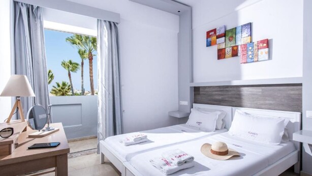 A modern hotel room with a bed, a desk with a lamp, a mirror, and a view of palm trees. - I Candy ''By Estia'' (Hersonissos)