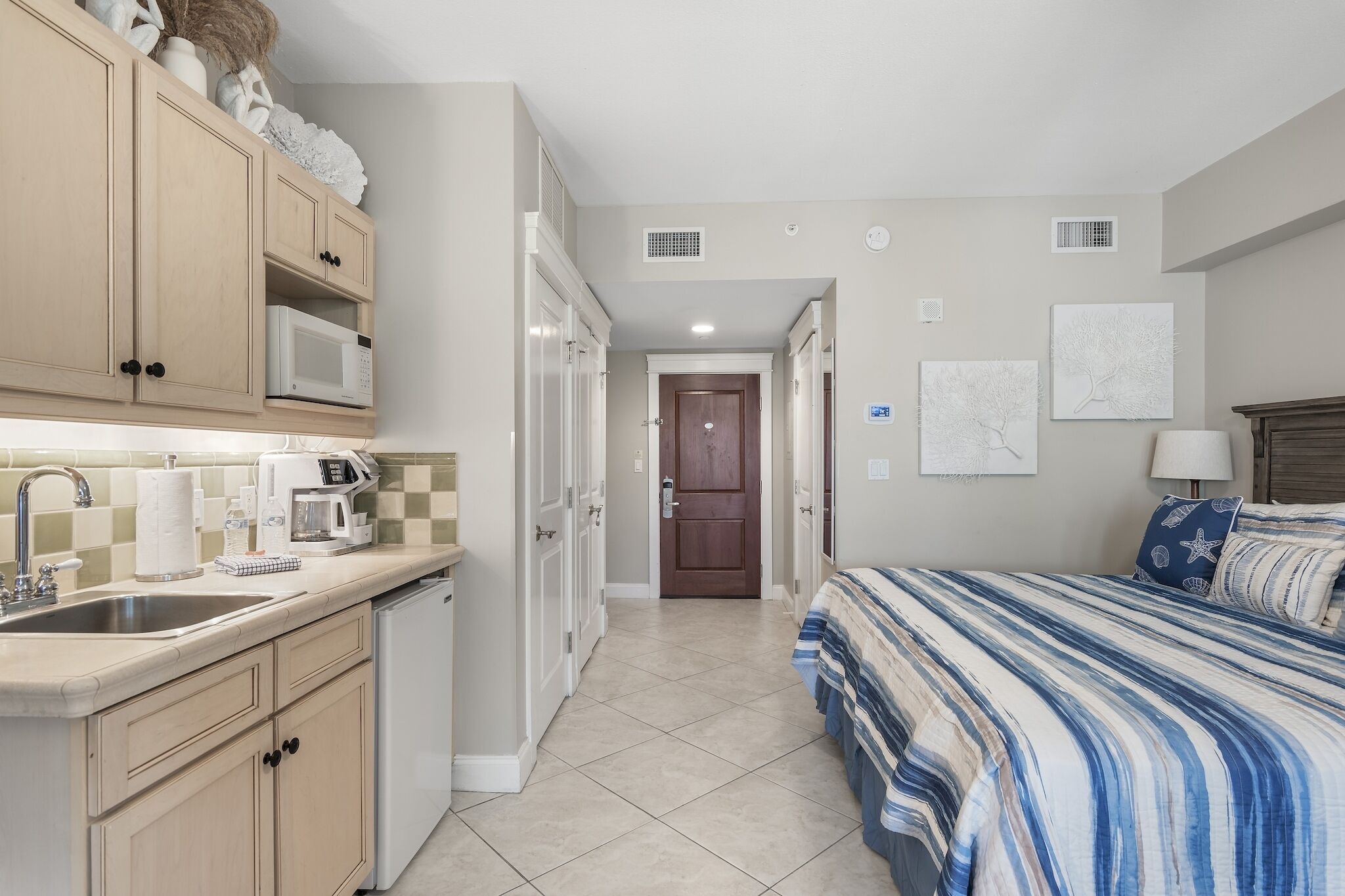 A compact kitchen with a microwave, coffee maker, and sink. A hallway leads to a bedroom with a bed and a nightstand.