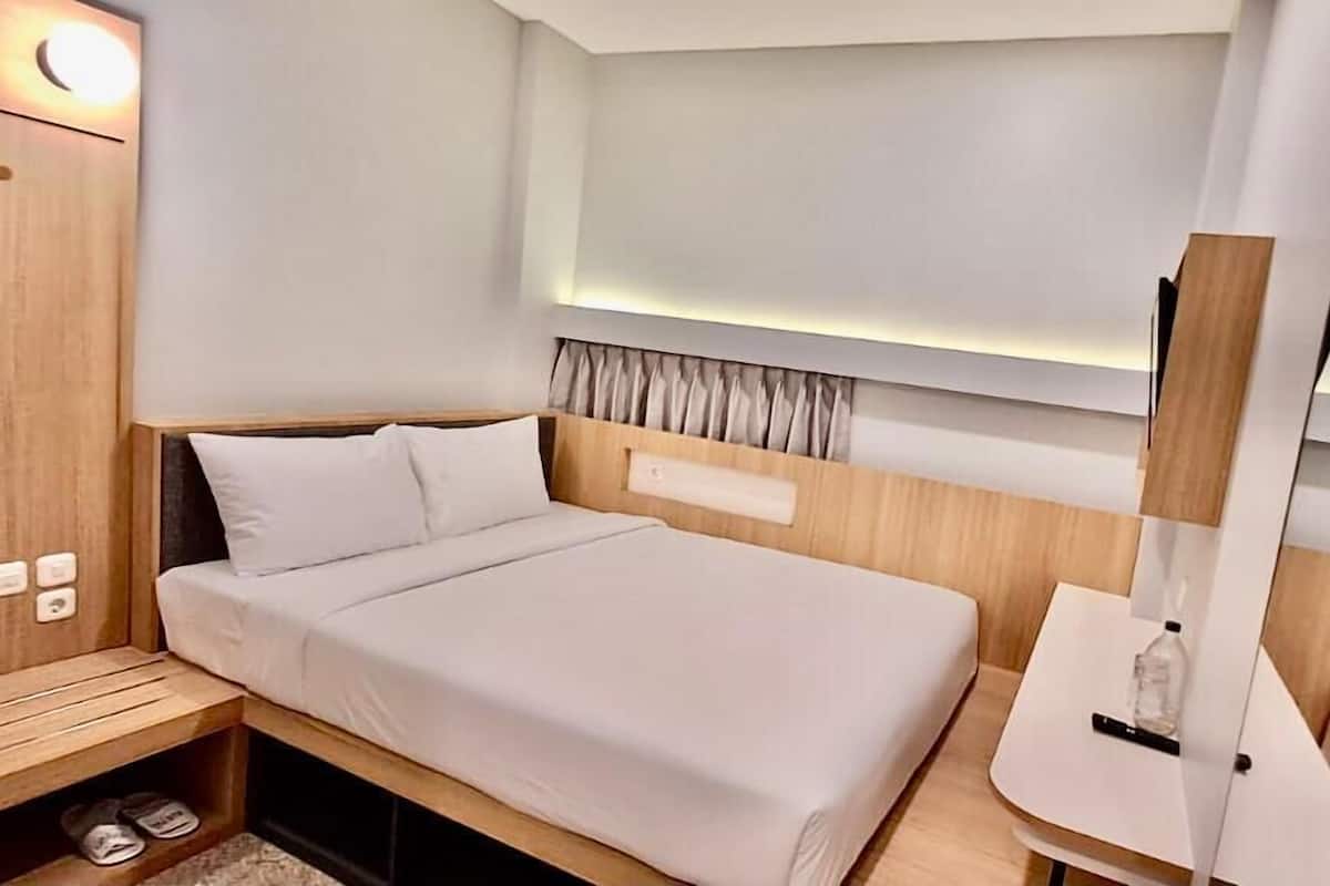 A modern hotel room with a neatly made bed, a wooden nightstand with slippers, a small table with a bottle, and a mirror.