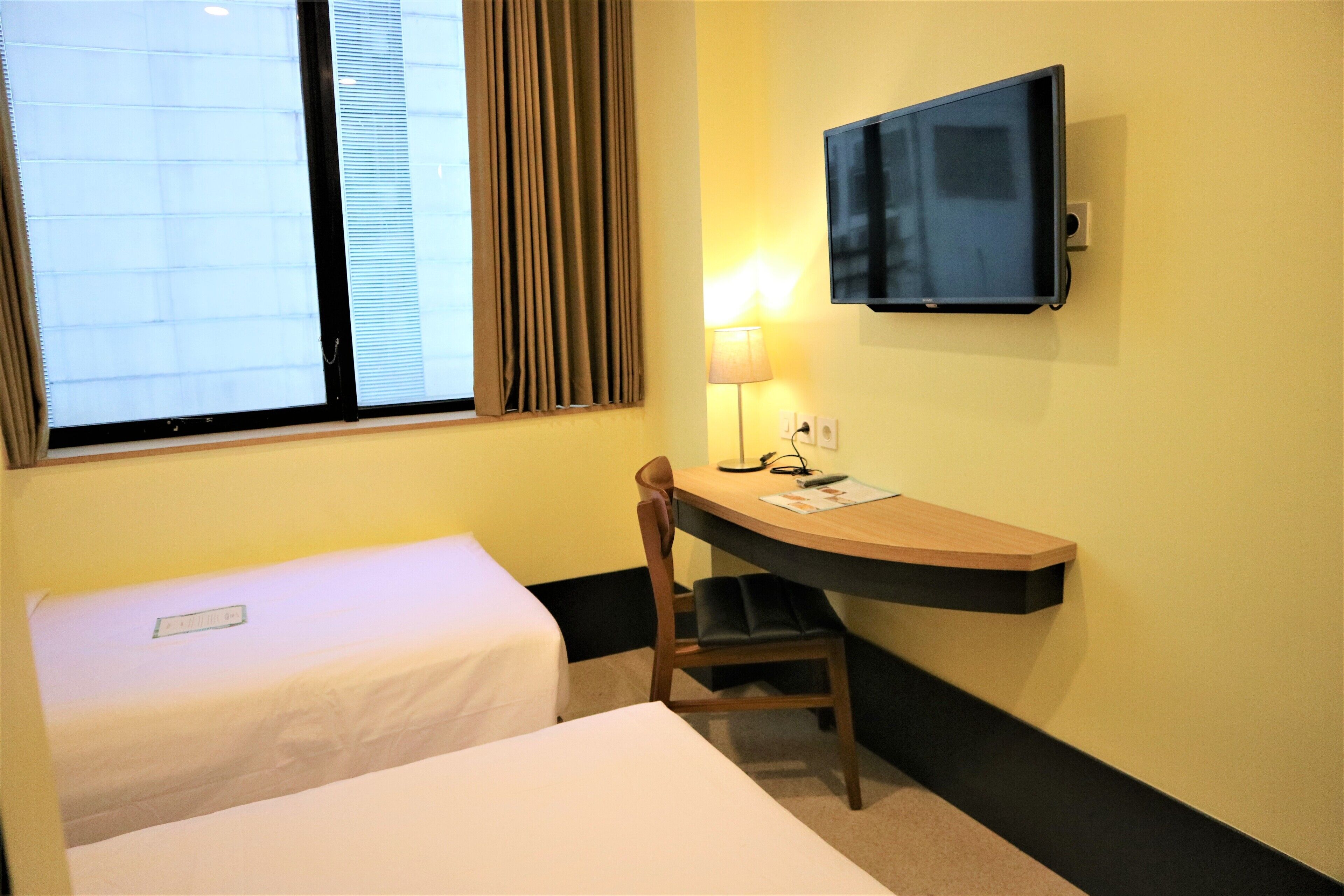Ein Hotelzimmer mit einem Einzelbett, einem Schreibtisch mit Stuhl, einem an der Wand befestigten Fernseher und einem Fenster mit Vorhängen.