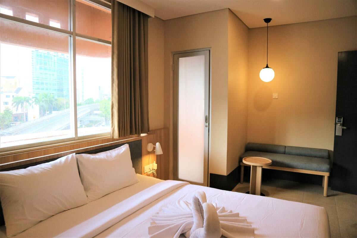 A hotel room with a bed, a bench, a small table, and a window with a view of buildings.