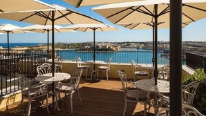 Een terras buiten met tafels en stoelen, grote parasols en uitzicht op een waterpartij en verre gebouwen.