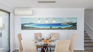 A dining area with a large painting of a coastal landscape, a glass table, and four chairs. - Pelican Sands 2 , 83 Soldiers Point Road - fantastic waterfront unit with pool and air conditioning (Soldiers Point)