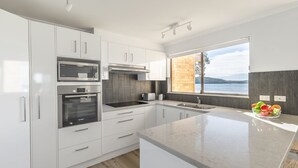 A modern kitchen with white cabinets, stainless steel appliances, and a large window offering a view of the water. - Pelican Sands 2 , 83 Soldiers Point Road - fantastic waterfront unit with pool and air conditioning (Soldiers Point)