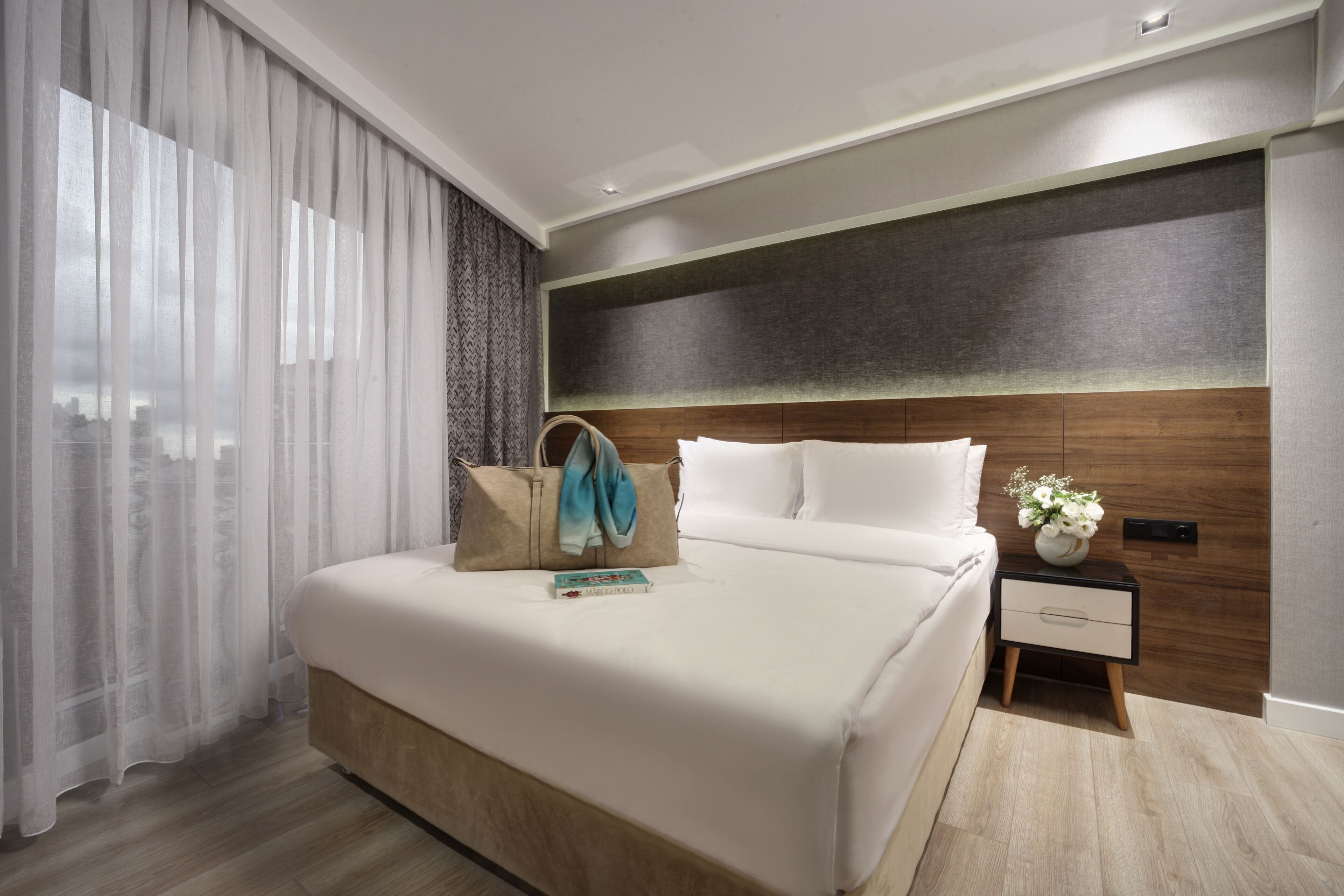 A modern hotel room with a large bed, a nightstand, a wooden headboard, and a window with sheer curtains.