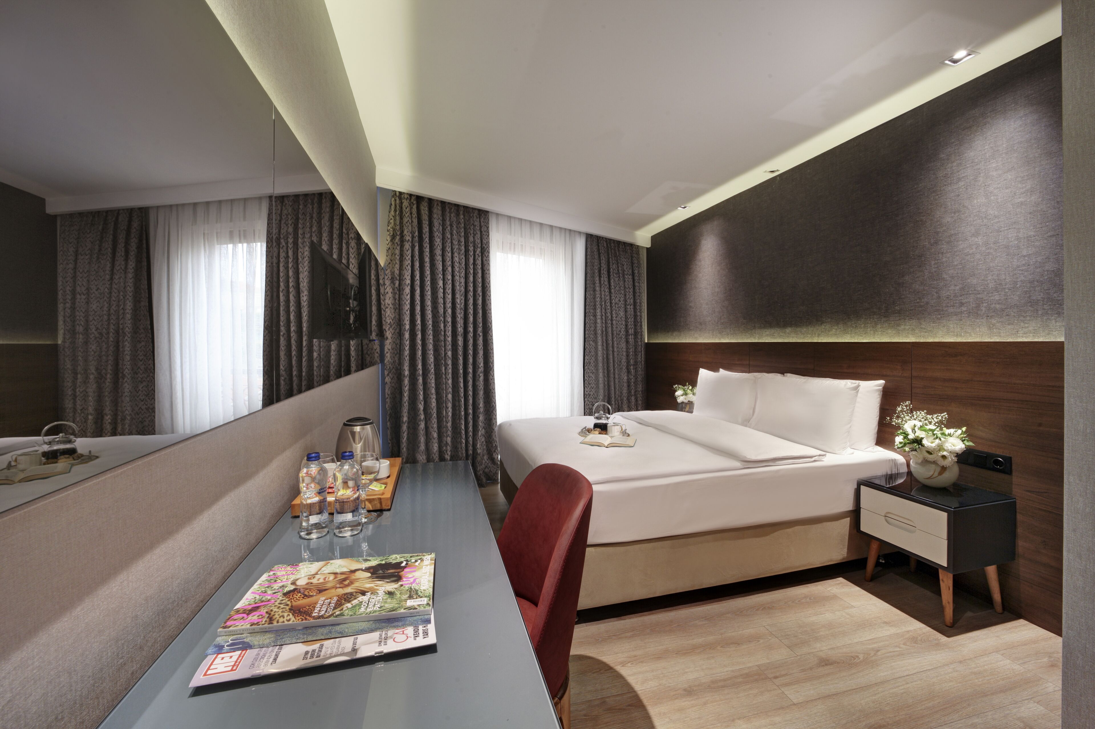 Une chambre d’hôtel moderne avec un lit soigneusement fait, un bureau avec un magazine, une chaise, une table de chevet avec un vase et un grand miroir.