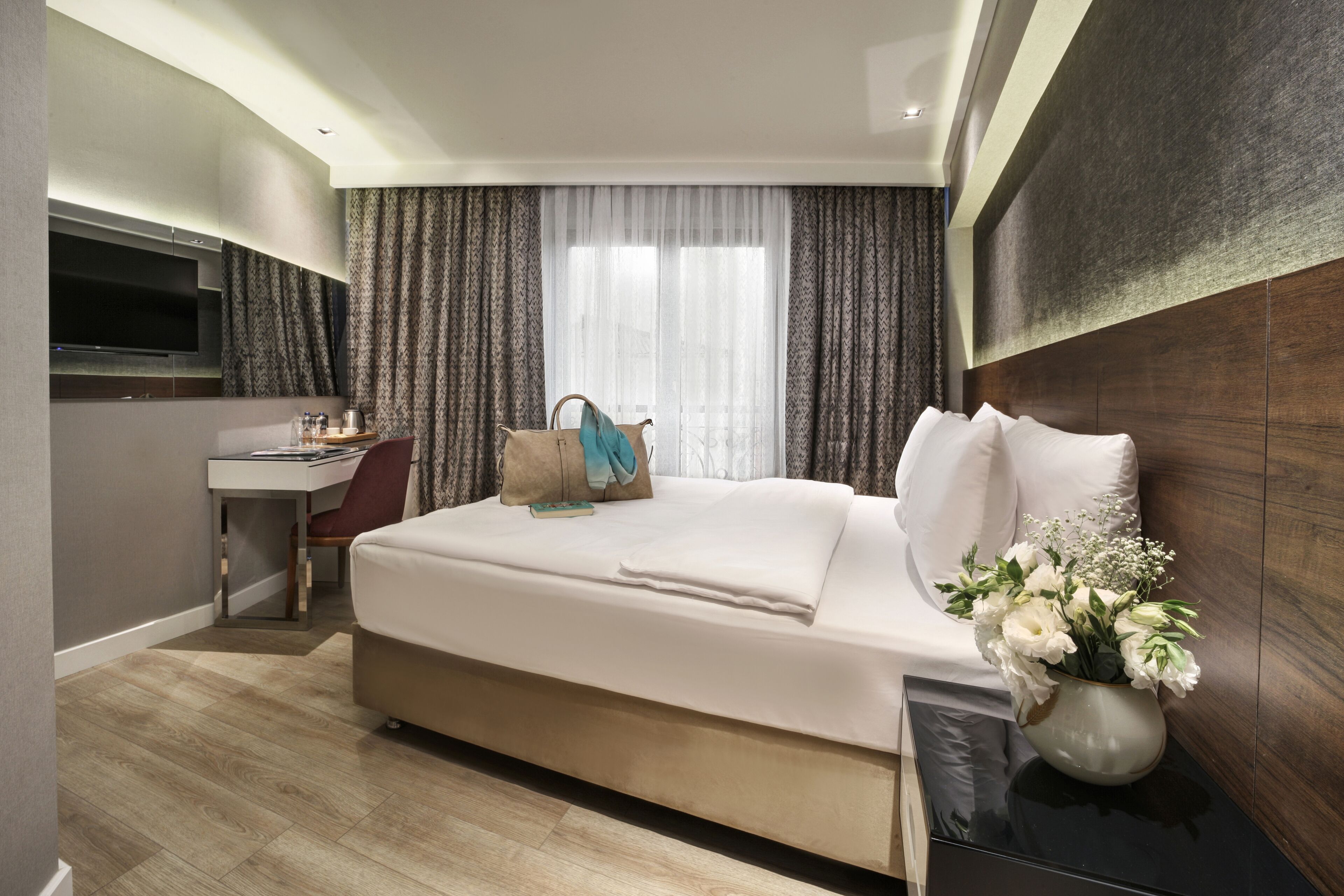 A modern hotel room with a bed, a desk, a TV, and a vase of flowers.