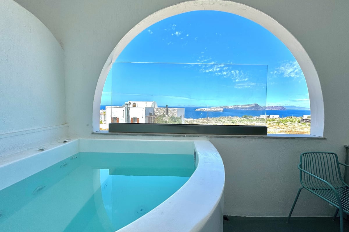 A room with a large arched window, a bathtub, and a view of a coastal landscape.