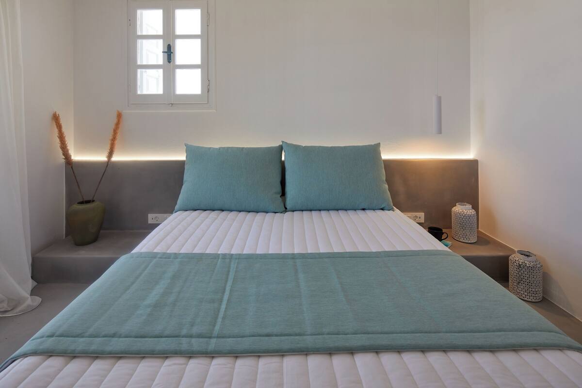 A neatly made bed with a teal bedspread and pillows, a bedside table with a lamp, and a window with a view.