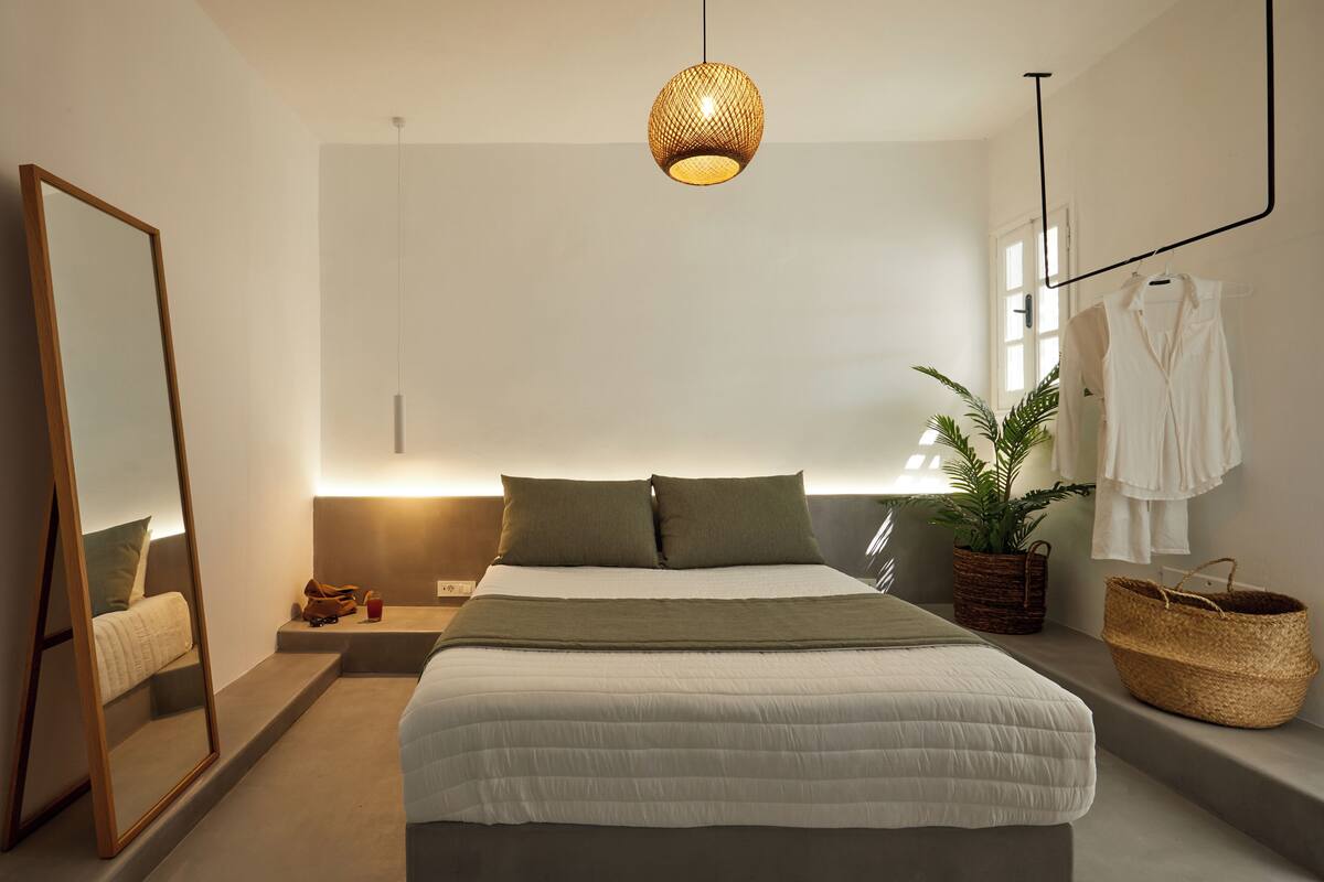 A modern bedroom with a bed, bedside table, mirror, hanging light fixture, window, and a potted plant.