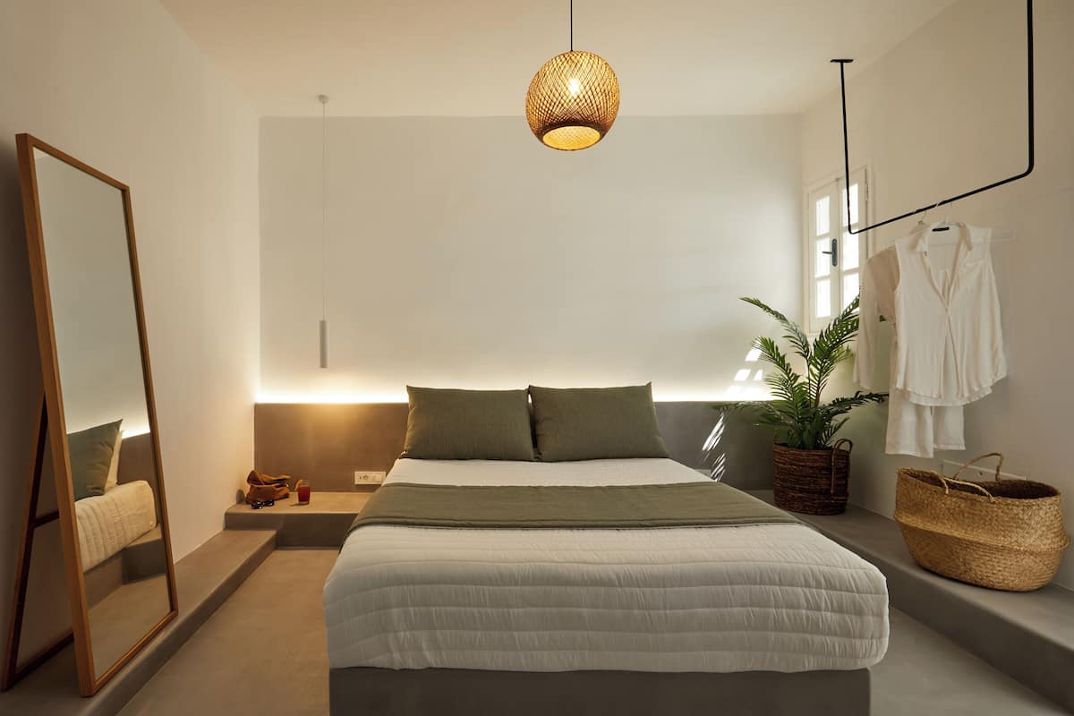 Ein modernes Schlafzimmer mit Bett, Nachttisch, Spiegel, Pendelleuchte, Fenster und einer Topfpflanze.