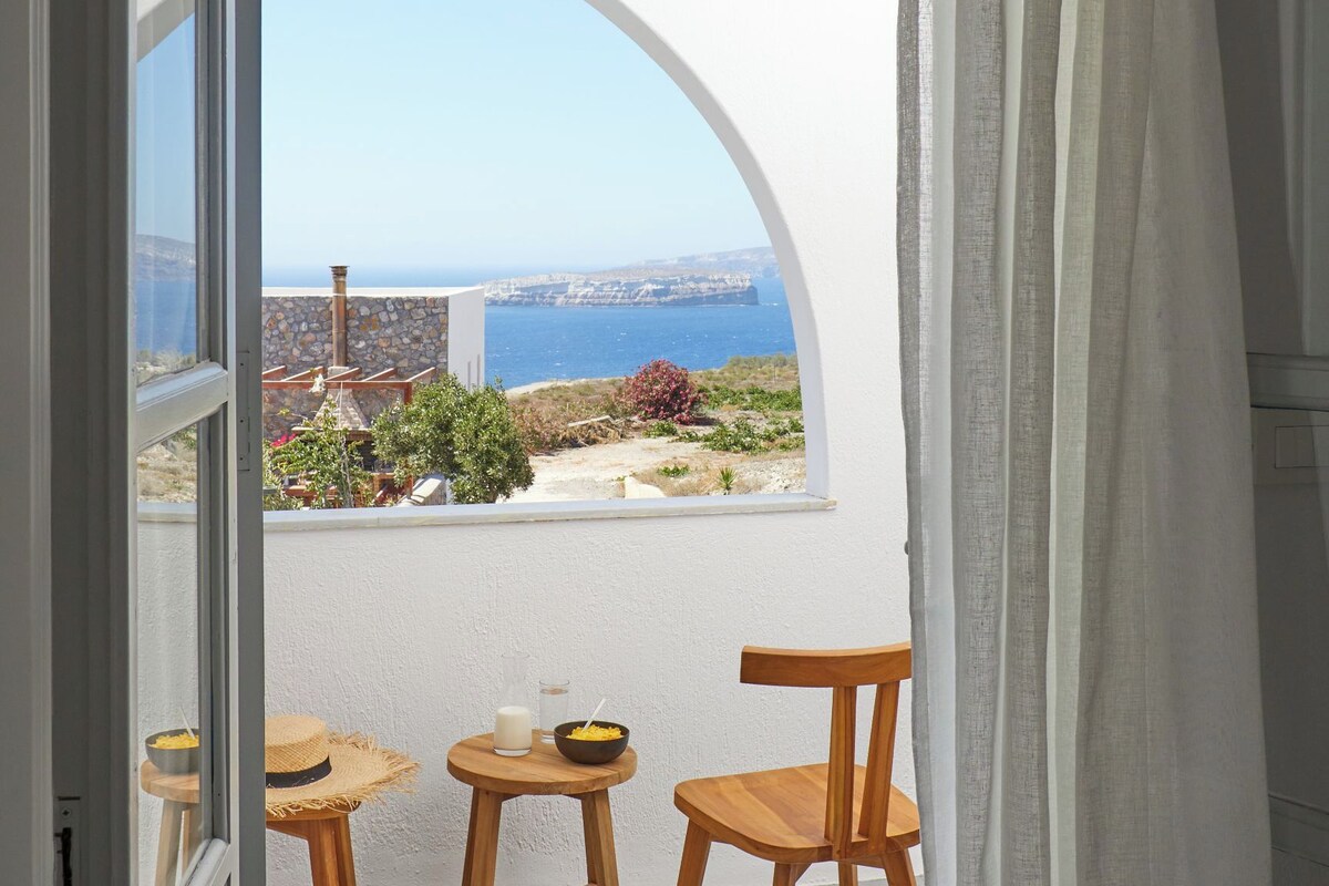 An open door leading to a balcony with a view of the sea, a straw hat, and a wooden chair.