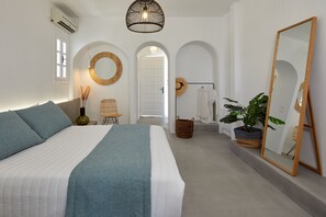 A bedroom with a bed, a large mirror, a plant, and a door leading to another room. - Nuvia Suites (Santorini)