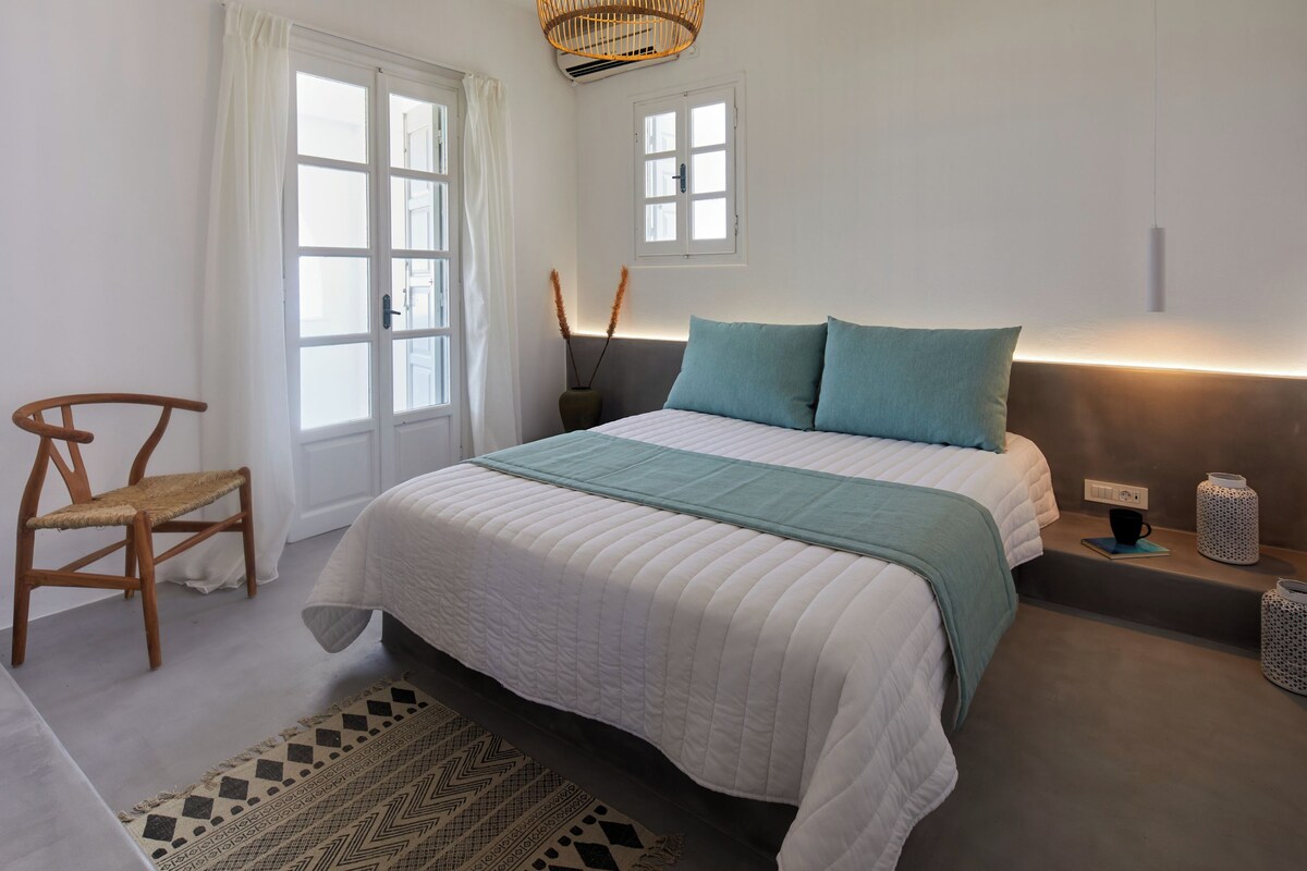 A bedroom with a bed, a chair, a window, and a bedside table with a lamp.