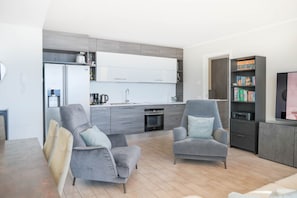 Un salon moderne avec deux fauteuils gris, une table à manger et une cuisine en arrière-plan.