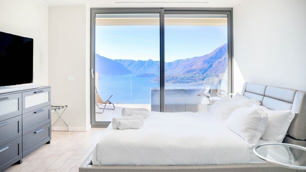 A modern bedroom with a large bed, a TV, and a view of a lake and mountains. - In a panoramic position, modern apartment on the first floor of a new residence with SPA. Large windows frame the unique landscape of Lake COMO.This apartment has a beautiful terrace, with table and chairs and a sofa. The living room is a large open spac (Perledo)