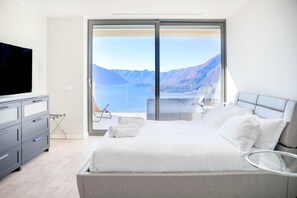 A modern bedroom with a large bed, a TV, and a view of a lake and mountains. - In a panoramic position, modern apartment on the first floor of a new residence with SPA. Large windows frame the unique landscape of Lake COMO.This apartment has a beautiful terrace, with table and chairs and a sofa. The living room is a large open spac (Perledo)