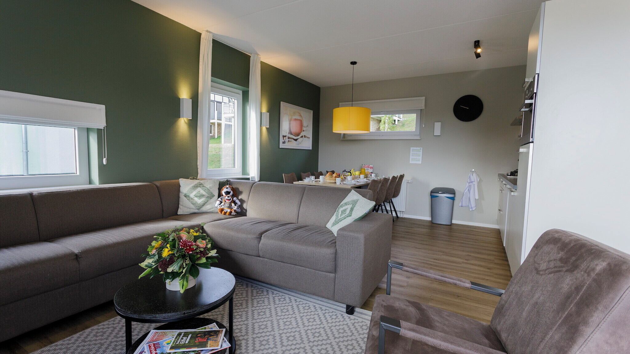 Een moderne woonkamer met een grijze bank, een klein rond tafeltje met een bloemenvaas, en een eethoek met stoelen en een gedekte tafel.