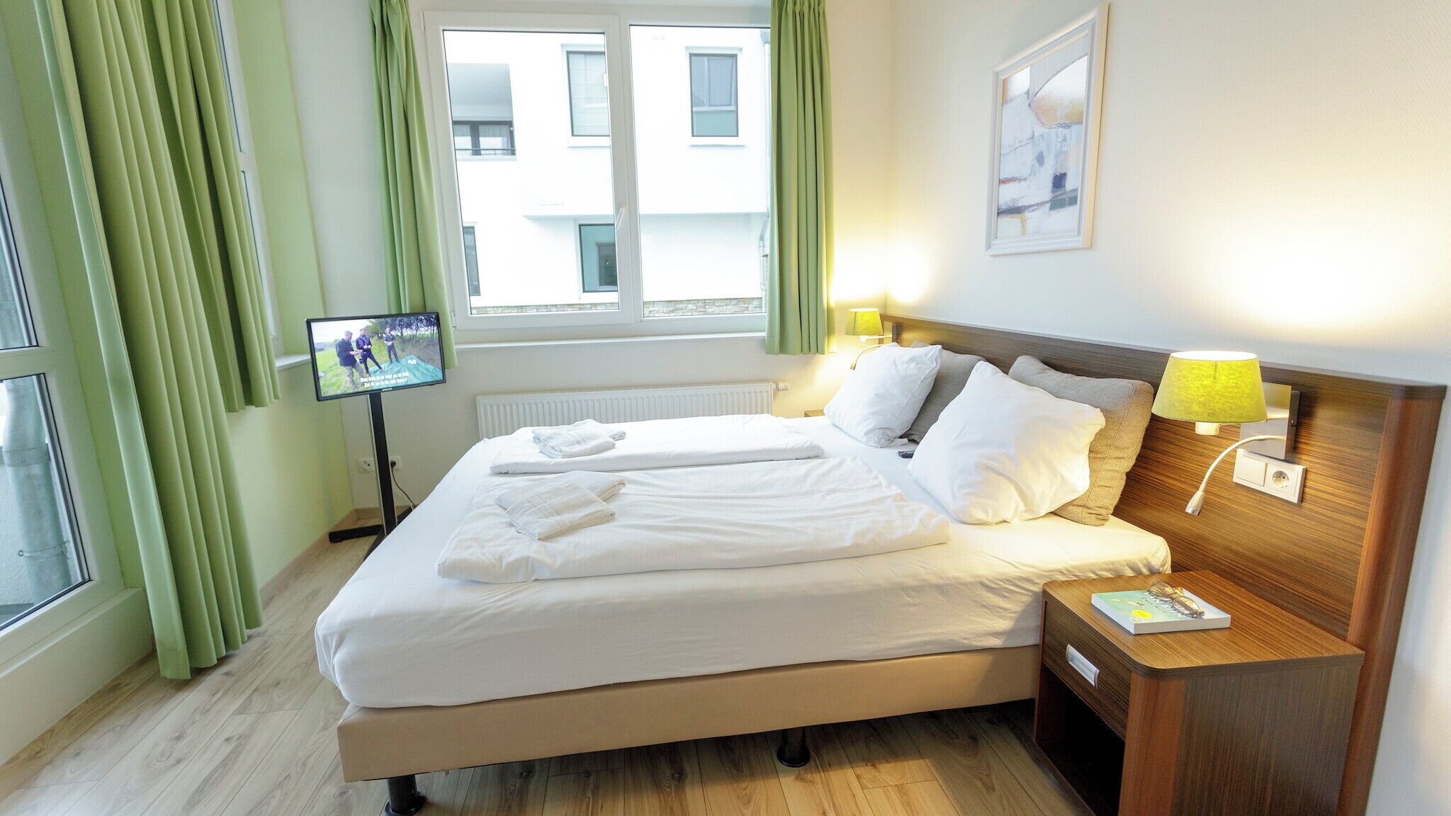 Ein Hotelzimmer mit Bett, Fernseher auf einem Schrank, Nachttisch mit Lampe und einem Fenster mit Vorhängen.