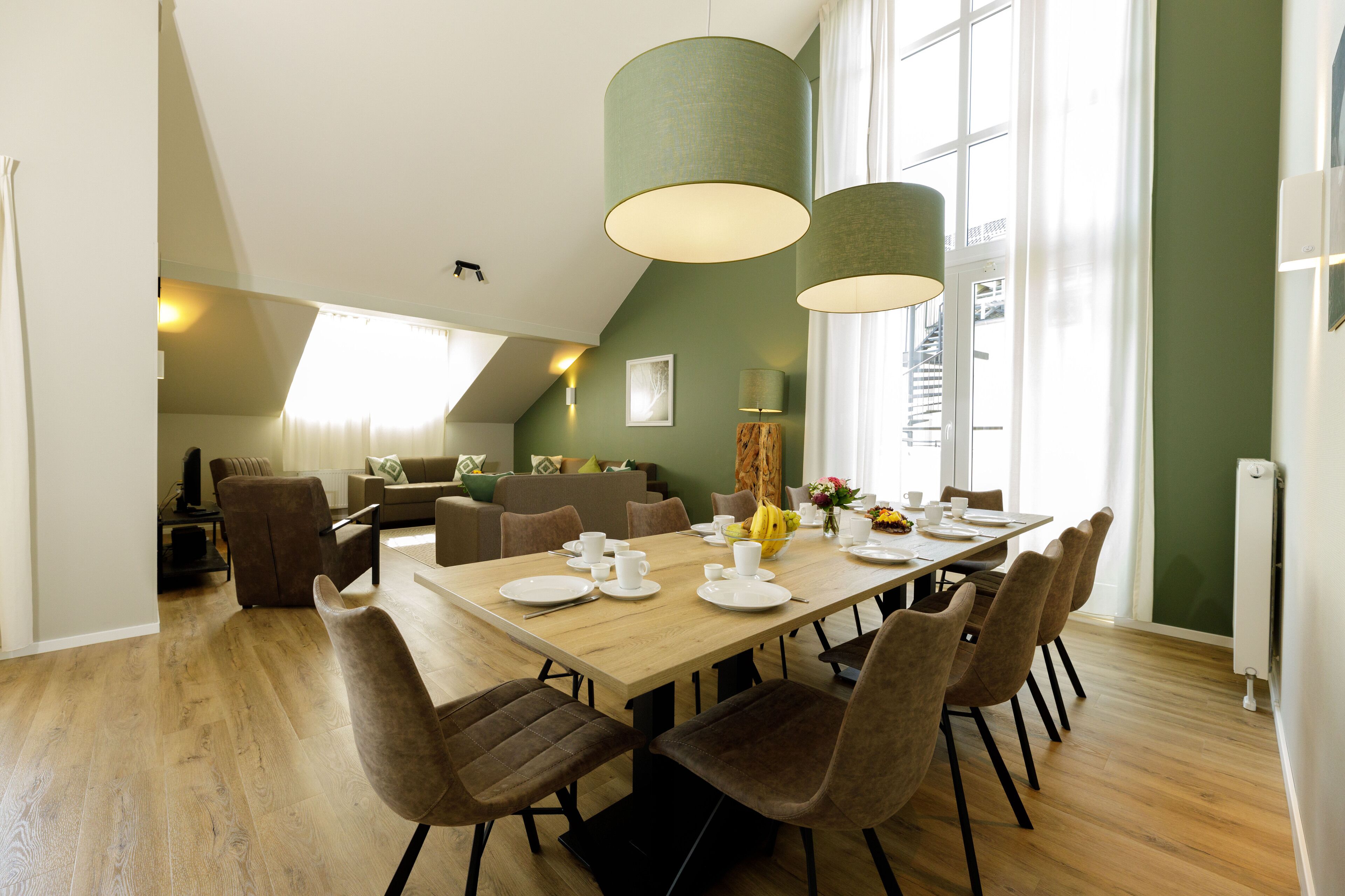 A dining area with a long table set for six, surrounded by six chairs. There are two hanging lamps above the table, and a window with curtains to the left.