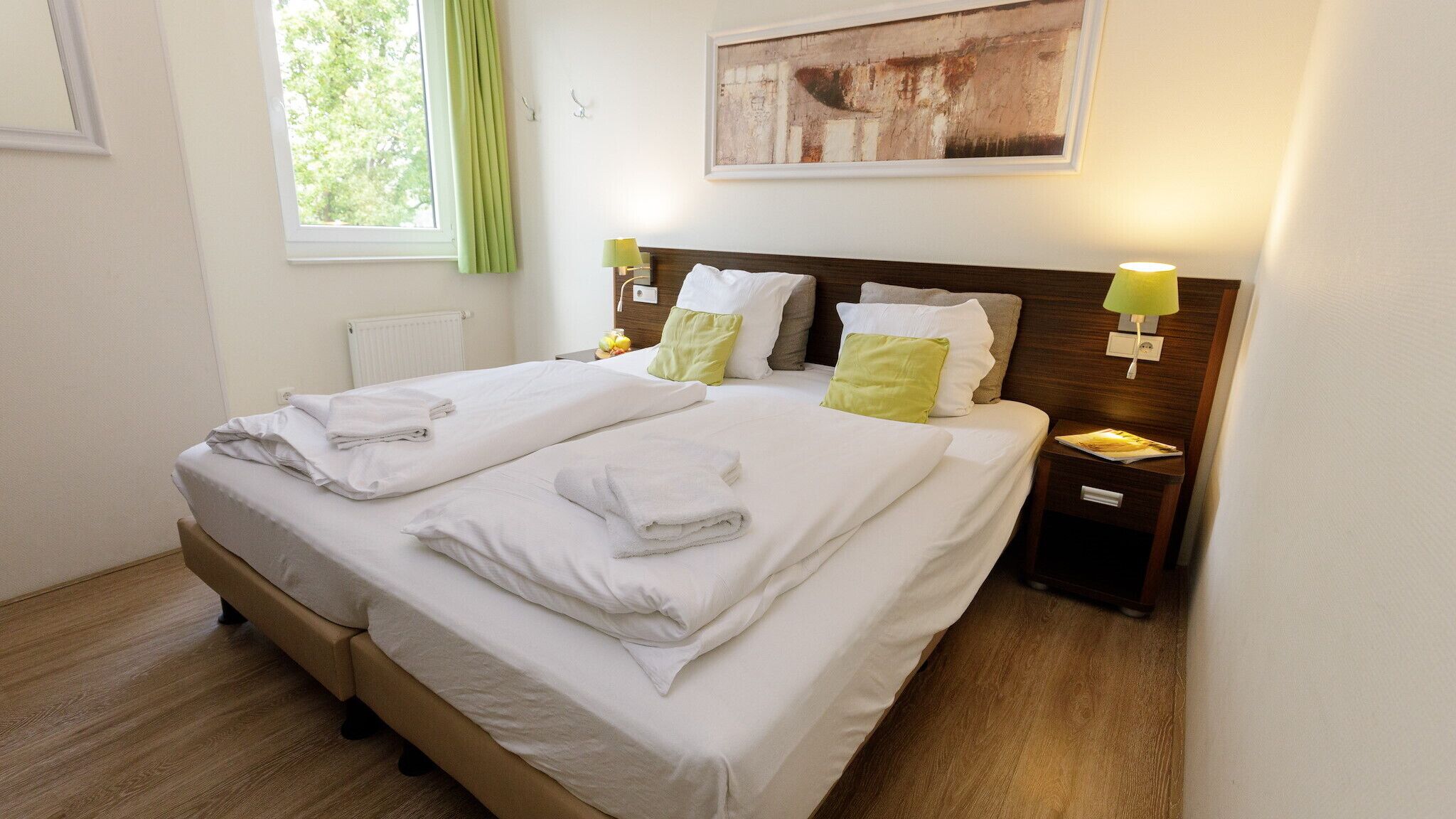 A neatly made double bed with white linens and towels, a wooden headboard, two bedside lamps, and a framed abstract painting on the wall.