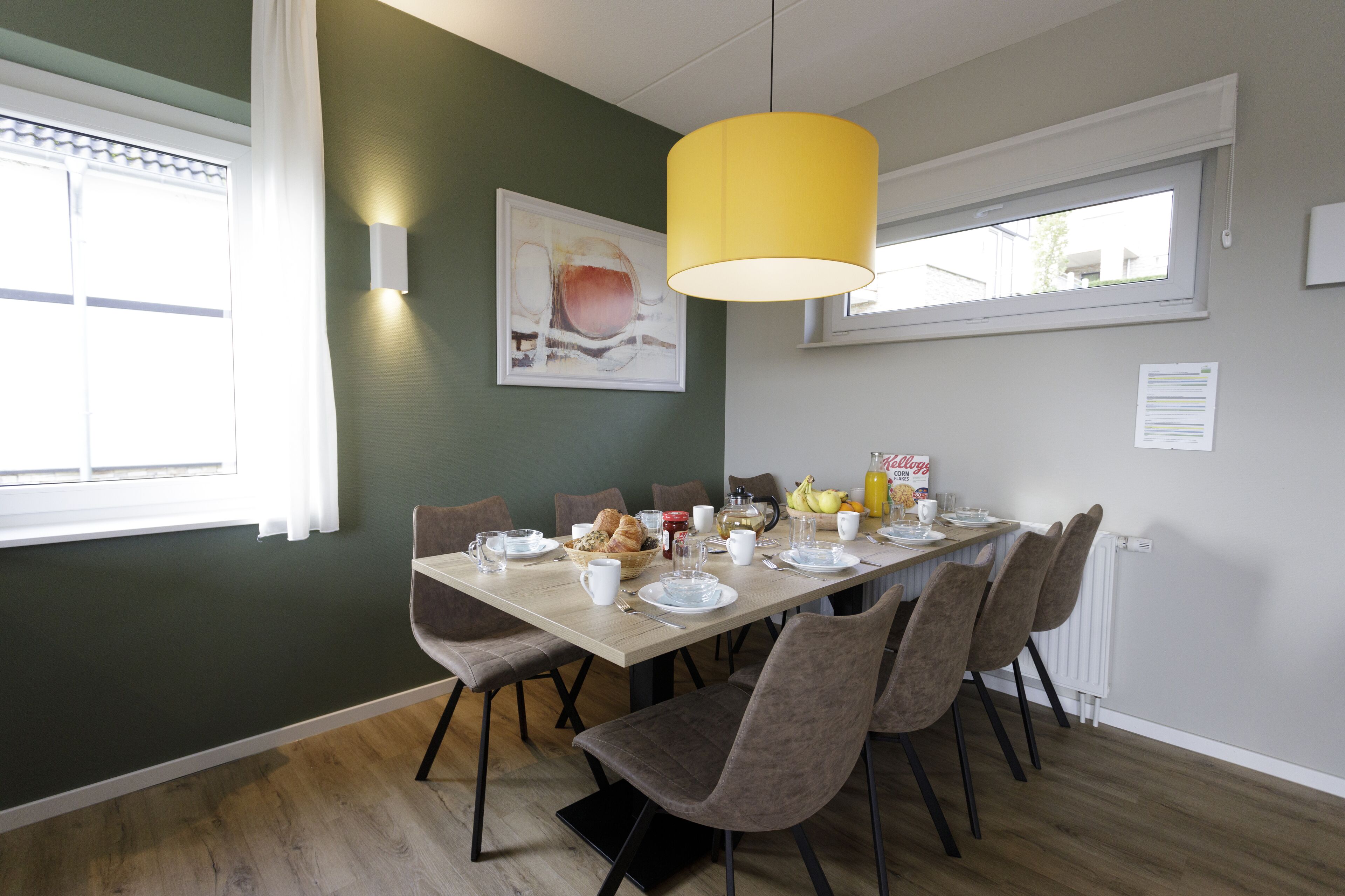 Een eethoek met een tafel voor vier personen, stoelen, een geel hanglamp, een raam met witte gordijnen en een schilderij aan de muur.