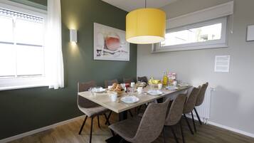 Een eethoek met een tafel voor vier personen, stoelen, een geel hanglamp, een raam met witte gordijnen en een schilderij aan de muur.
