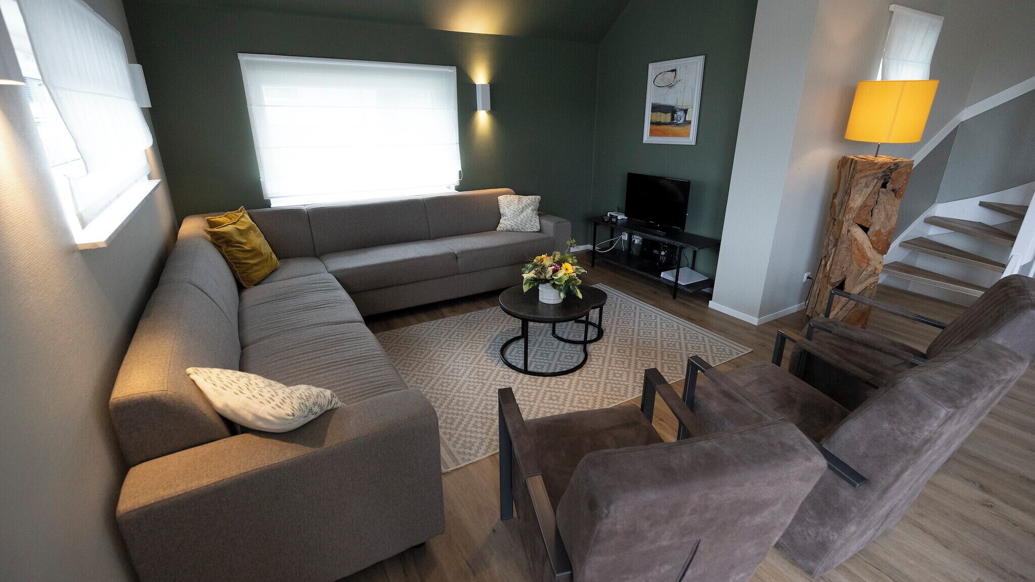 En moderne stue med en grå sofa, et sofabord med blomster, et fjernsyn og en trappe.