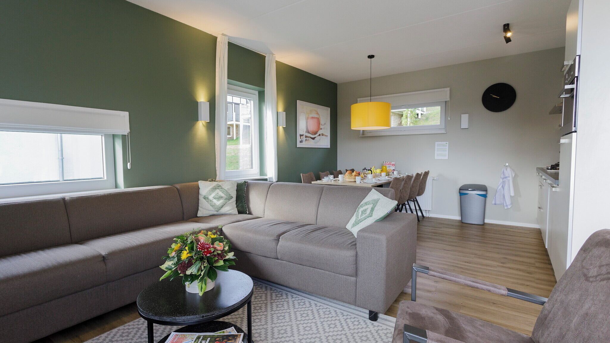 A modern living room with a sofa, a coffee table with flowers, a dining area with chairs and a table, and a kitchen area with a refrigerator.