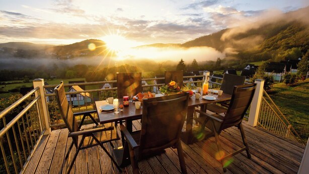 A wooden deck with a table set for breakfast overlooking a mountainous landscape at sunrise. - Dormio resort Eifeler Tor (Heimbach)