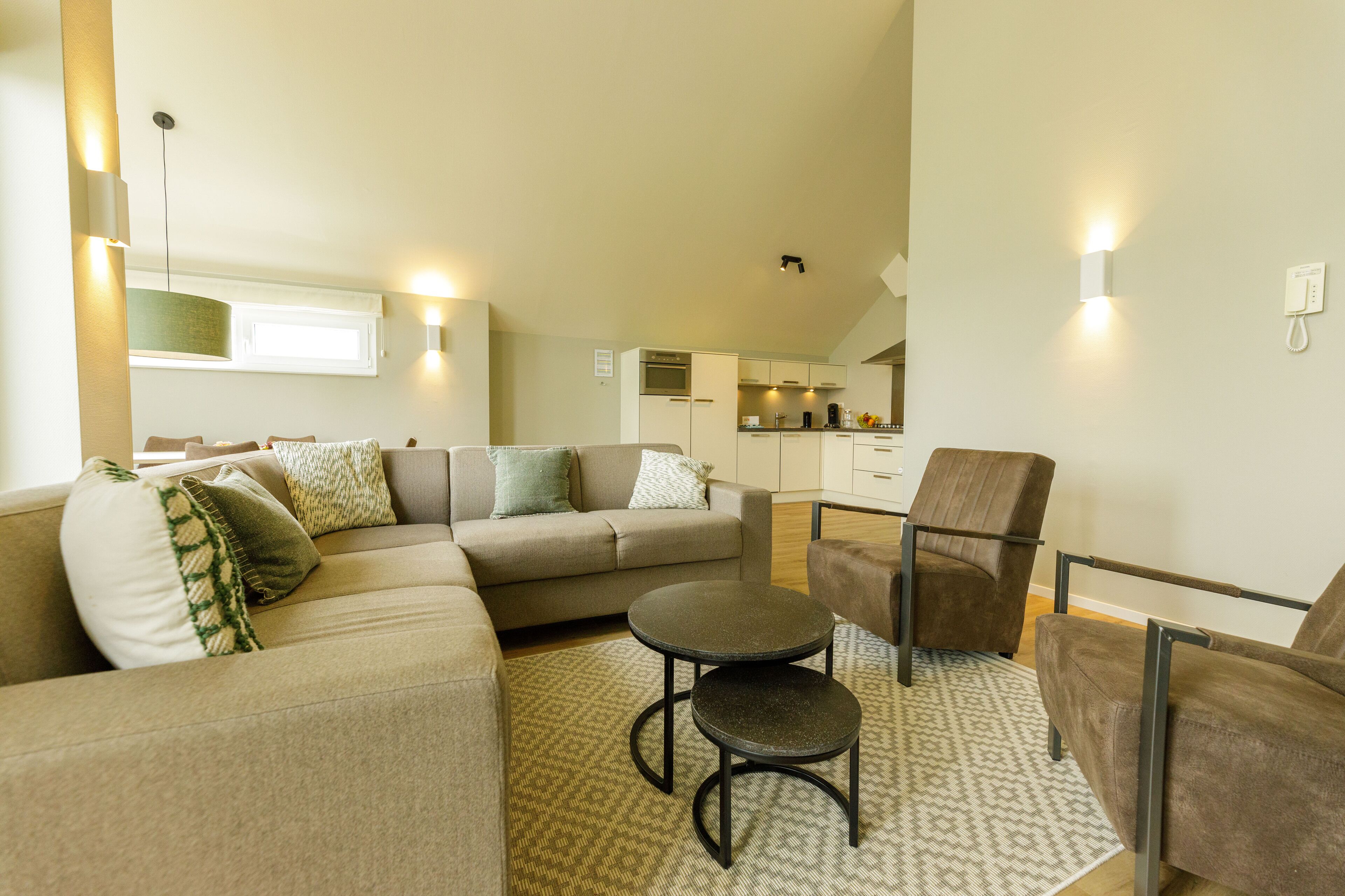 A modern living room with a sectional sofa, a round coffee table, and a brown armchair. A kitchen is visible in the background.