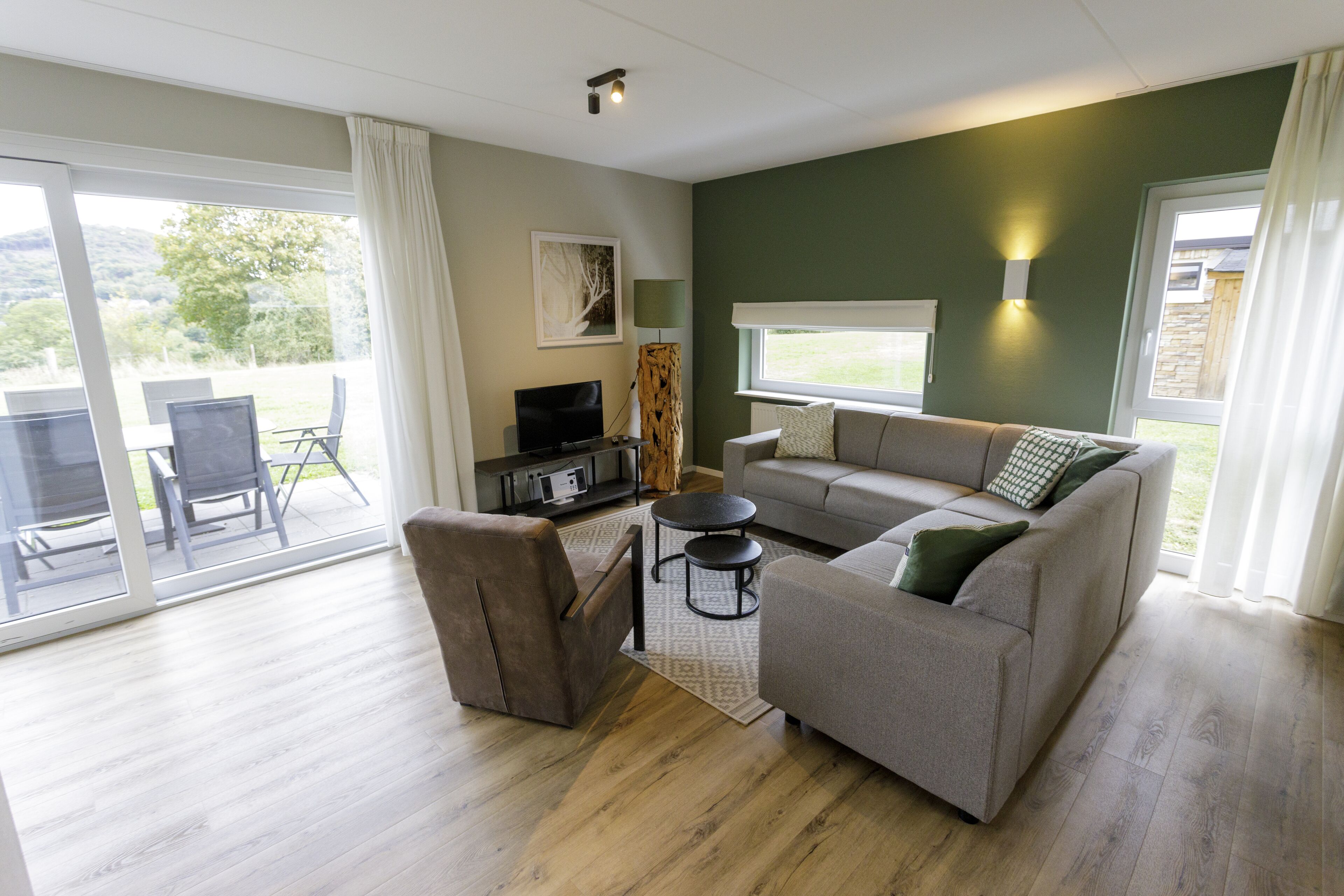 A modern living room with a grey sofa, a brown armchair, and a TV stand. There is a large sliding glass door leading to an outdoor patio area.
