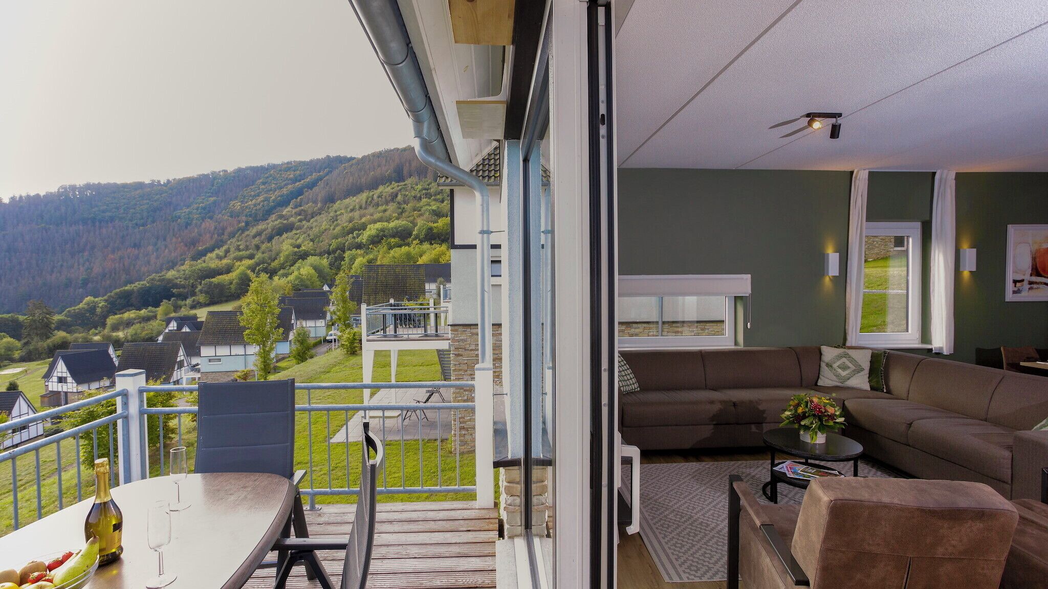 Un balcone con vista su un borgo collinare, un divano e un tavolo da pranzo con bicchieri di vino e frutta.