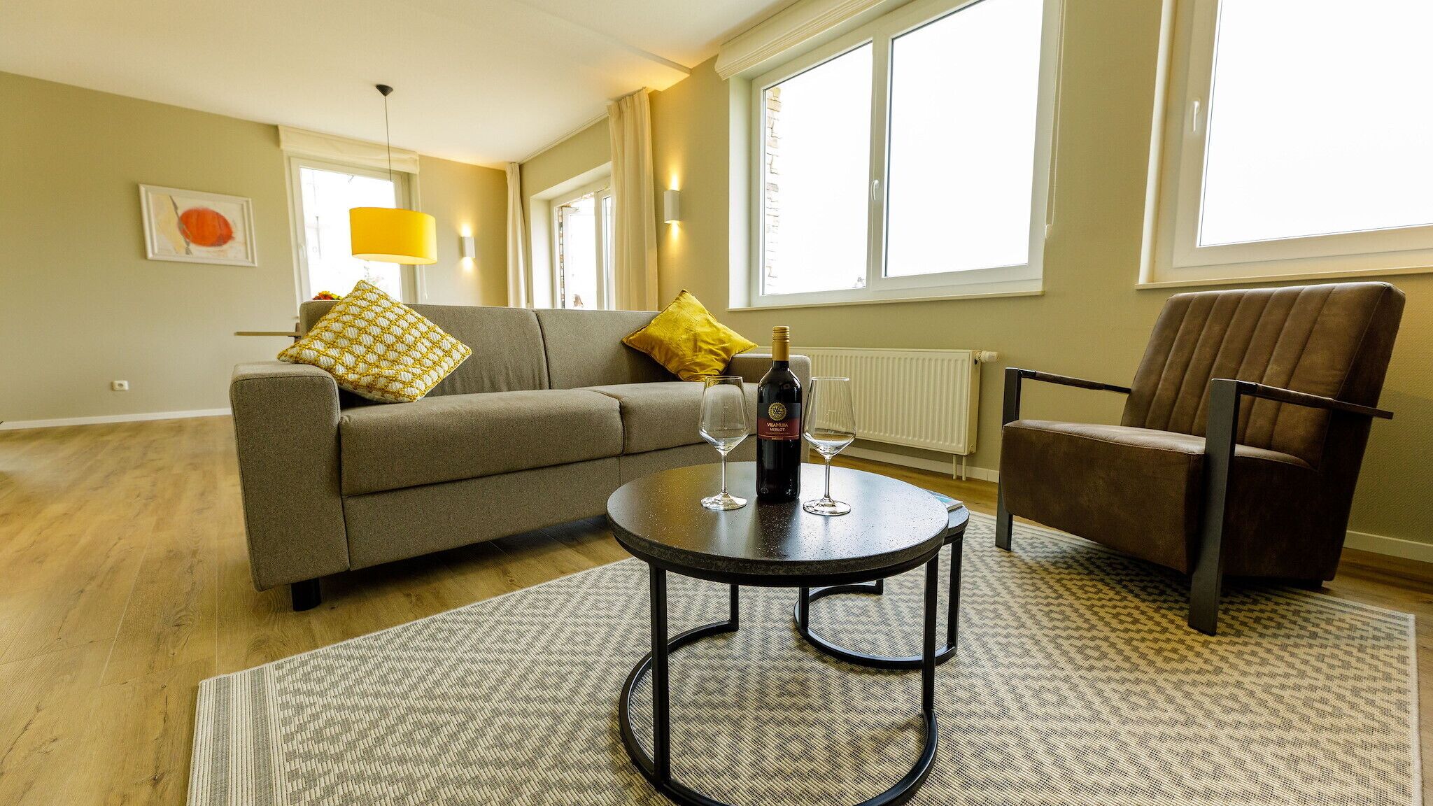 A living room with a sofa, armchair, round coffee table, and a bottle of wine.