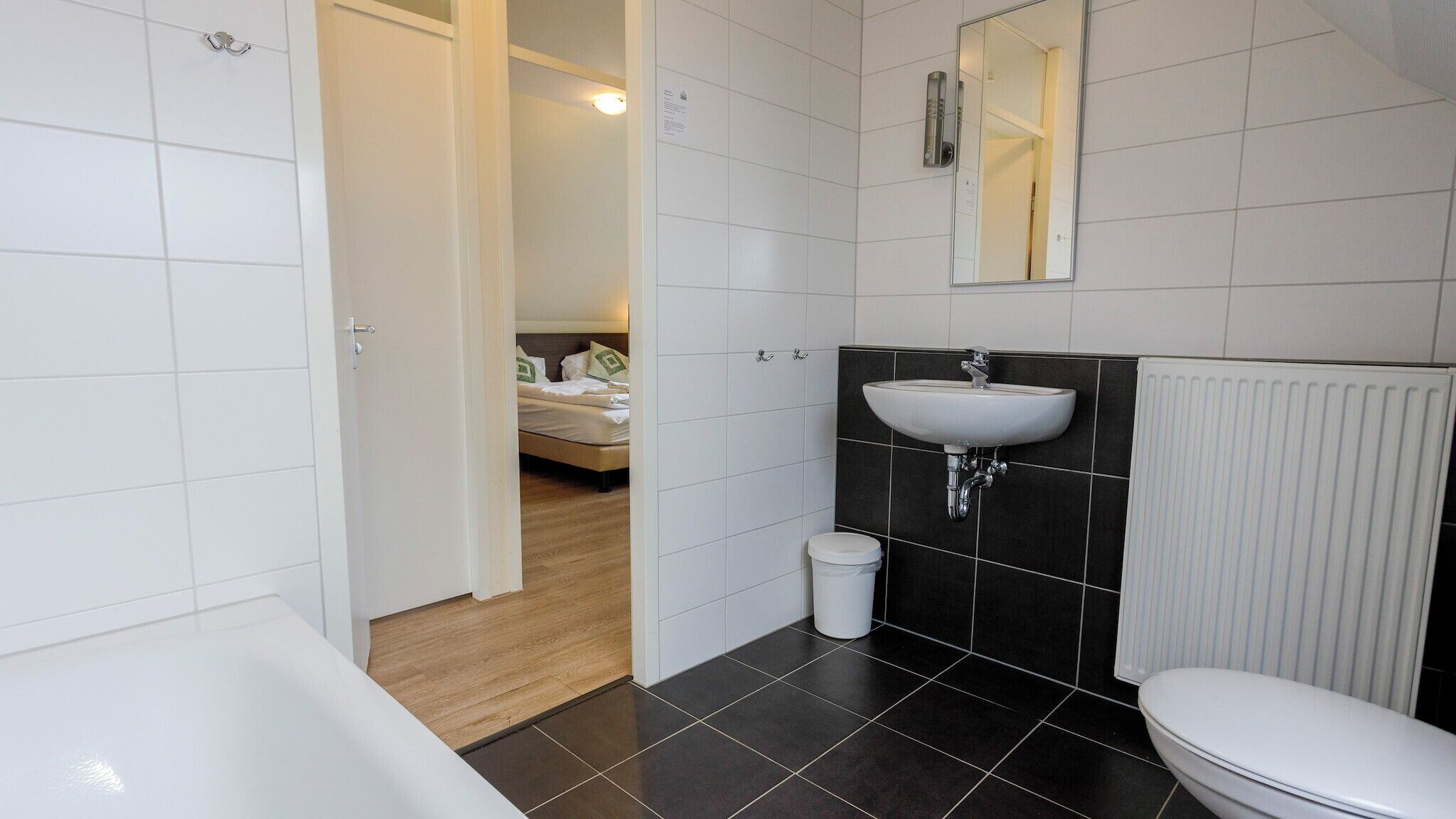 A modern bathroom with a bathtub, a sink, and a toilet. There is a wooden floor and a door leading to a bedroom.