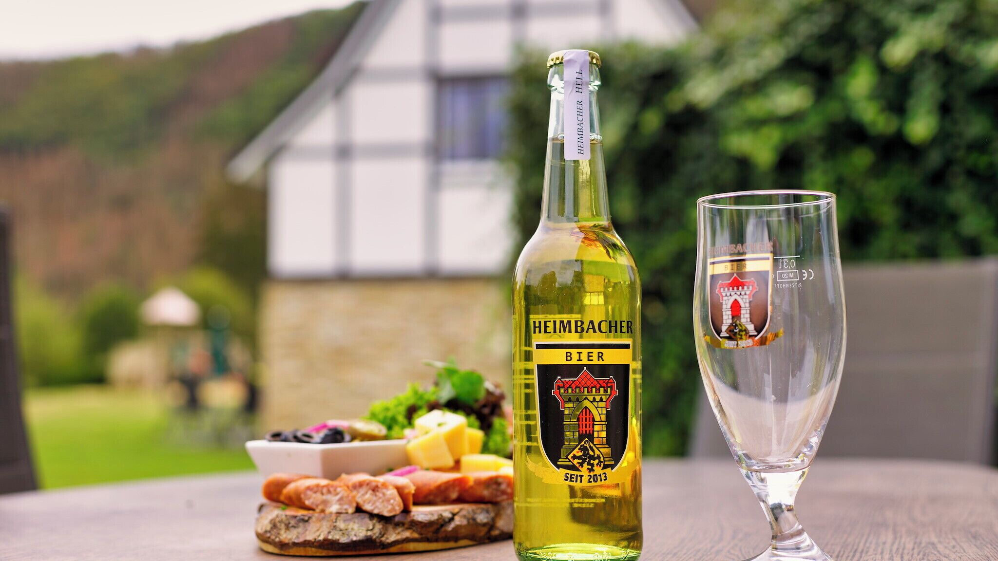 A bottle of Heimbacher Bier and a glass on a table with a variety of food items.