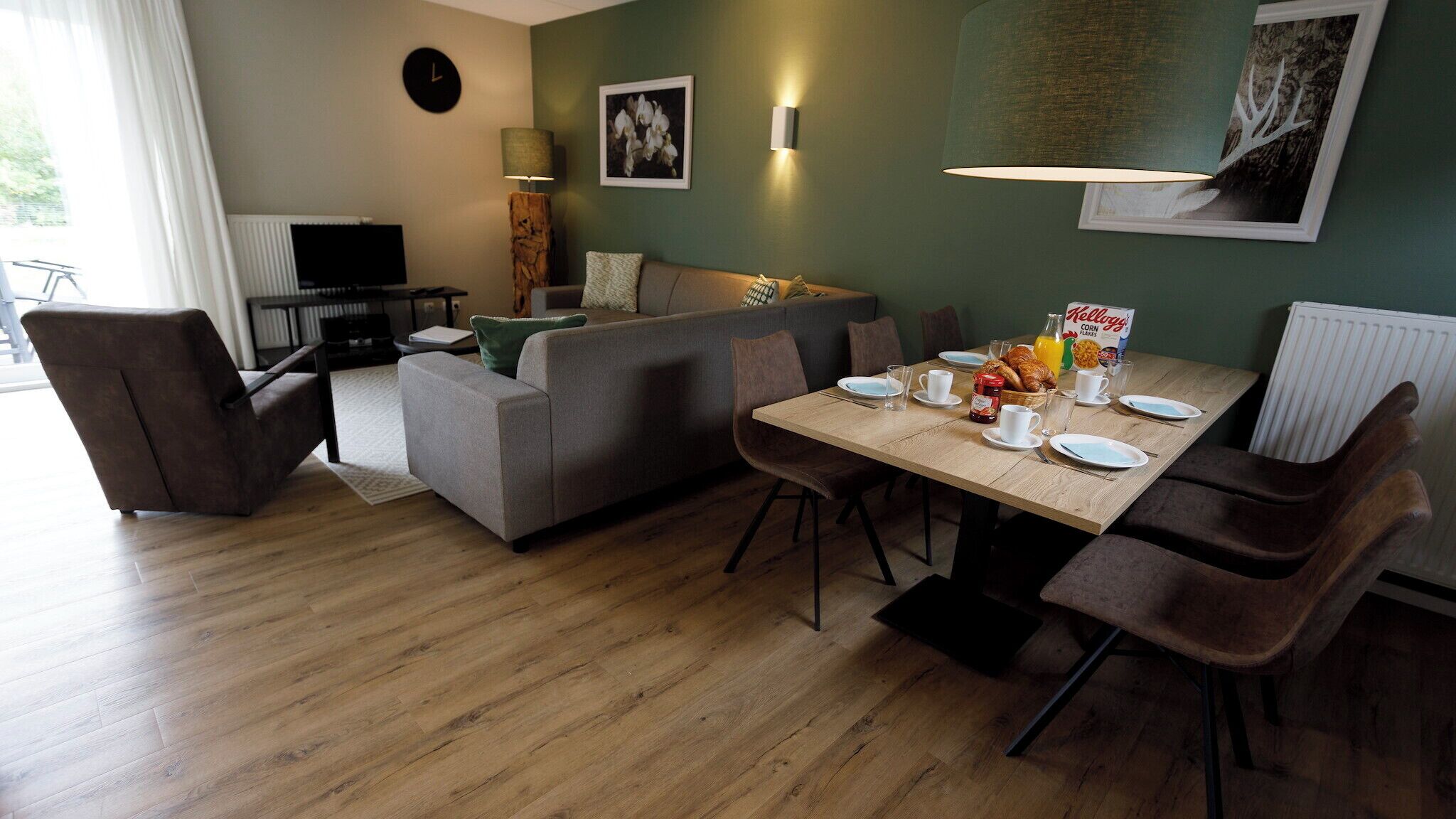 Een moderne woonkamer met een grijze bank, een eettafel gedekt voor het ontbijt, en een televisie in de hoek.