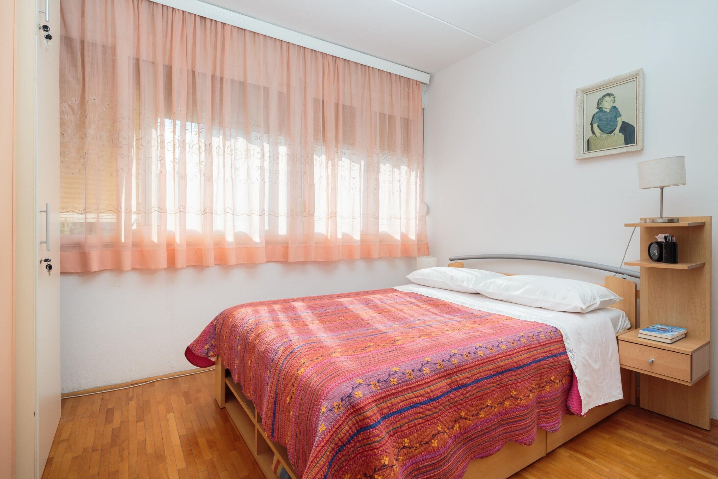 Une chambre à coucher avec un lit, une table de chevet avec une lampe, une étagère avec des livres et une fenêtre avec des rideaux légers.