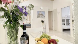 A dining area with a white table, a vase of flowers, a bottle of wine, and a bowl of fruit.