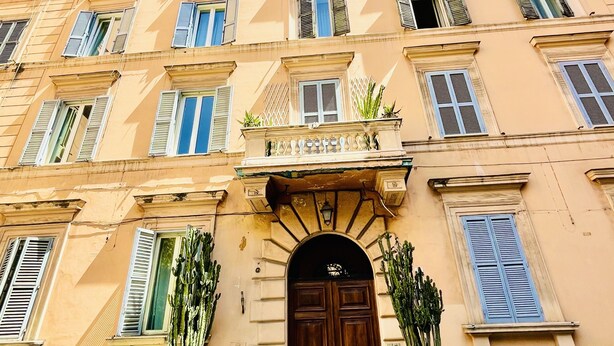 A multi-story building with a central entrance, large windows, and a balcony with potted plants.