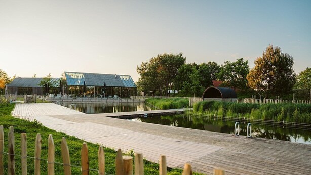 Een houten looppad dat naar een gebouw met glazen dak loopt, langs een gracht en een hek.