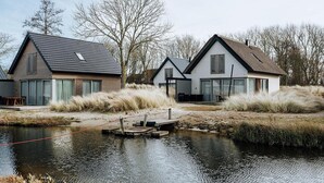 Twee moderne huizen met grote ramen, een steiger en een waterpartij ervoor.