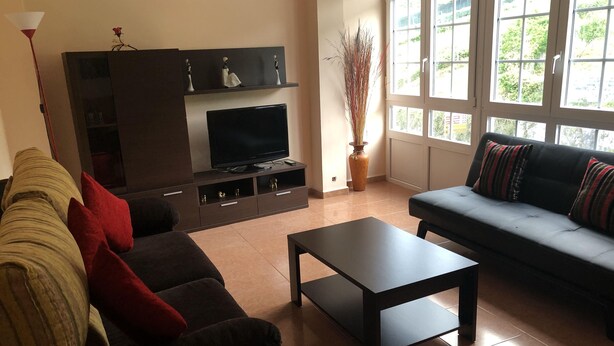 Una sala de estar con un sofá oscuro, un sofá rojo y beige, una mesa de centro negra, un televisor sobre un mueble de madera y ventanas amplias con vista a follaje.