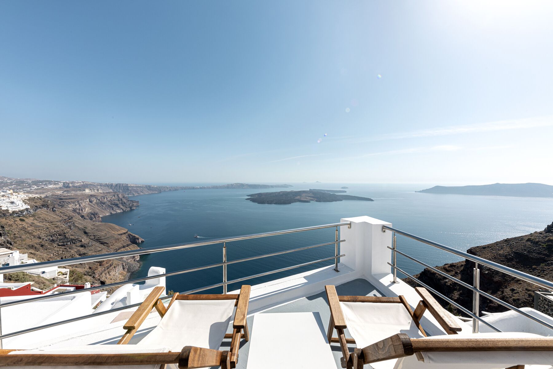 A balcony with wooden chairs and a white railing overlooking a coastal landscape with cliffs and a body of water.