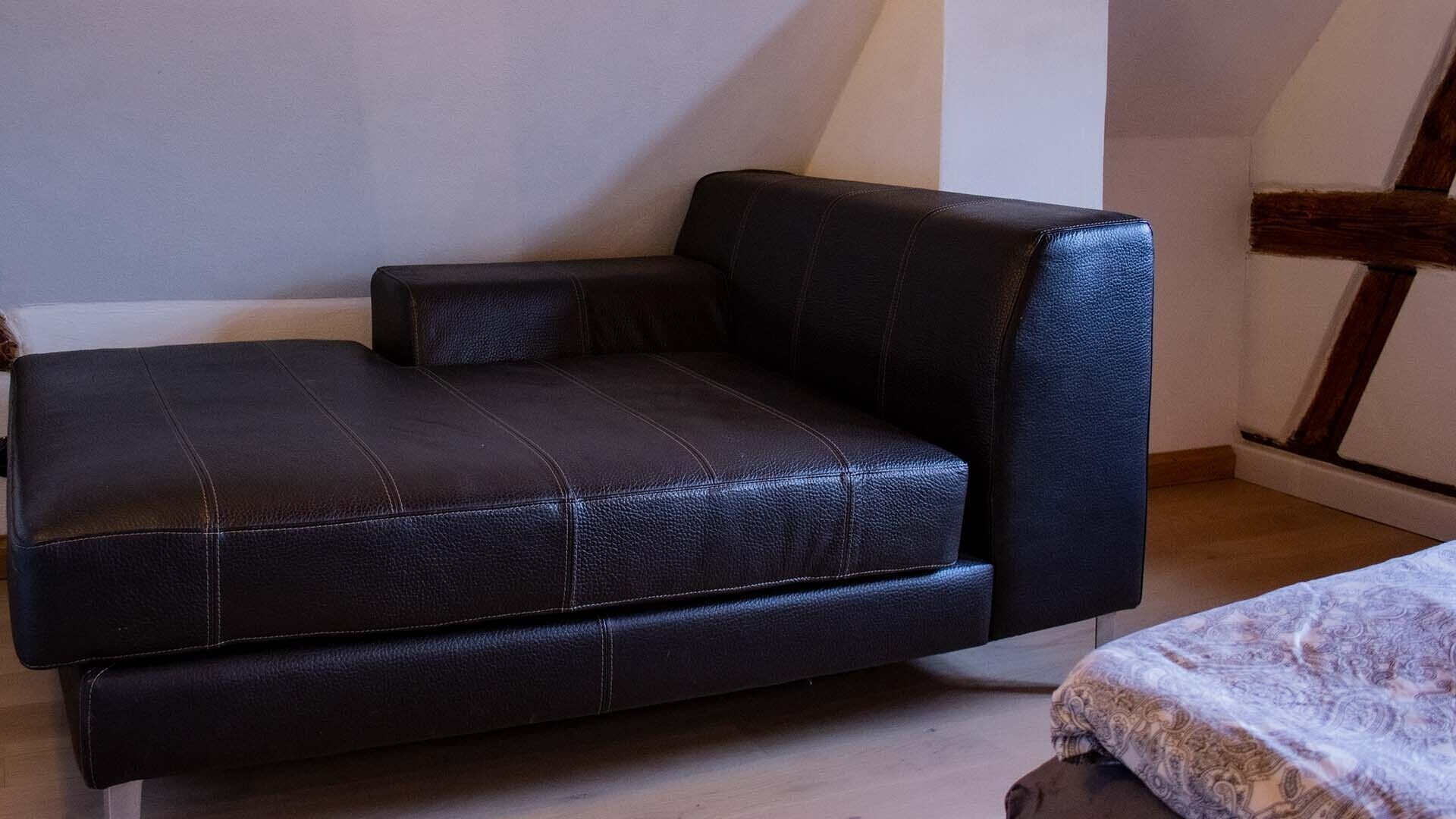 Un canapé en cuir noir, placé dans un coin d’une pièce dont le sol est en bois et les murs sont blancs.