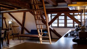 Une pièce rustique avec des poutres apparentes, un lit en mezzanine, un bureau avec une lampe et une vue sur l’extérieur grâce à de grandes fenêtres.