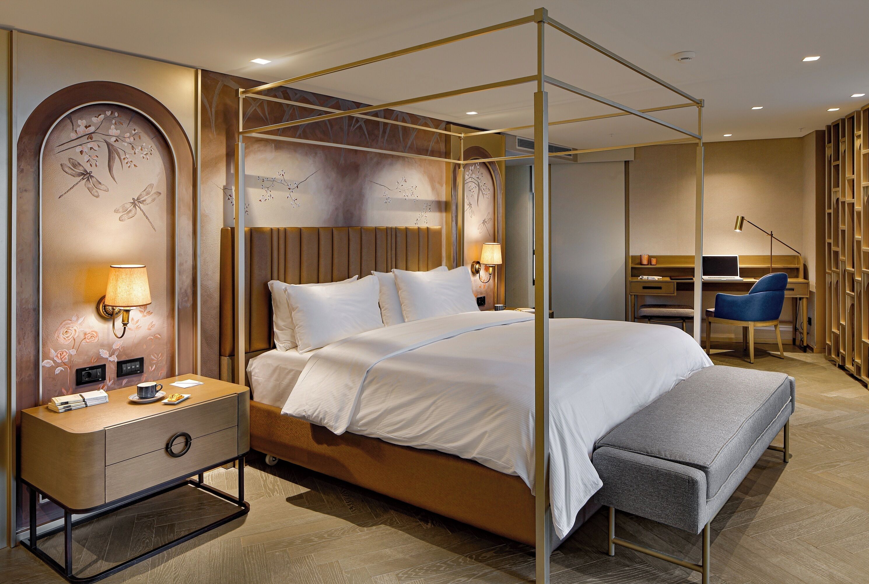 A modern hotel room with a four-poster bed, a desk with a chair, and a bookshelf.