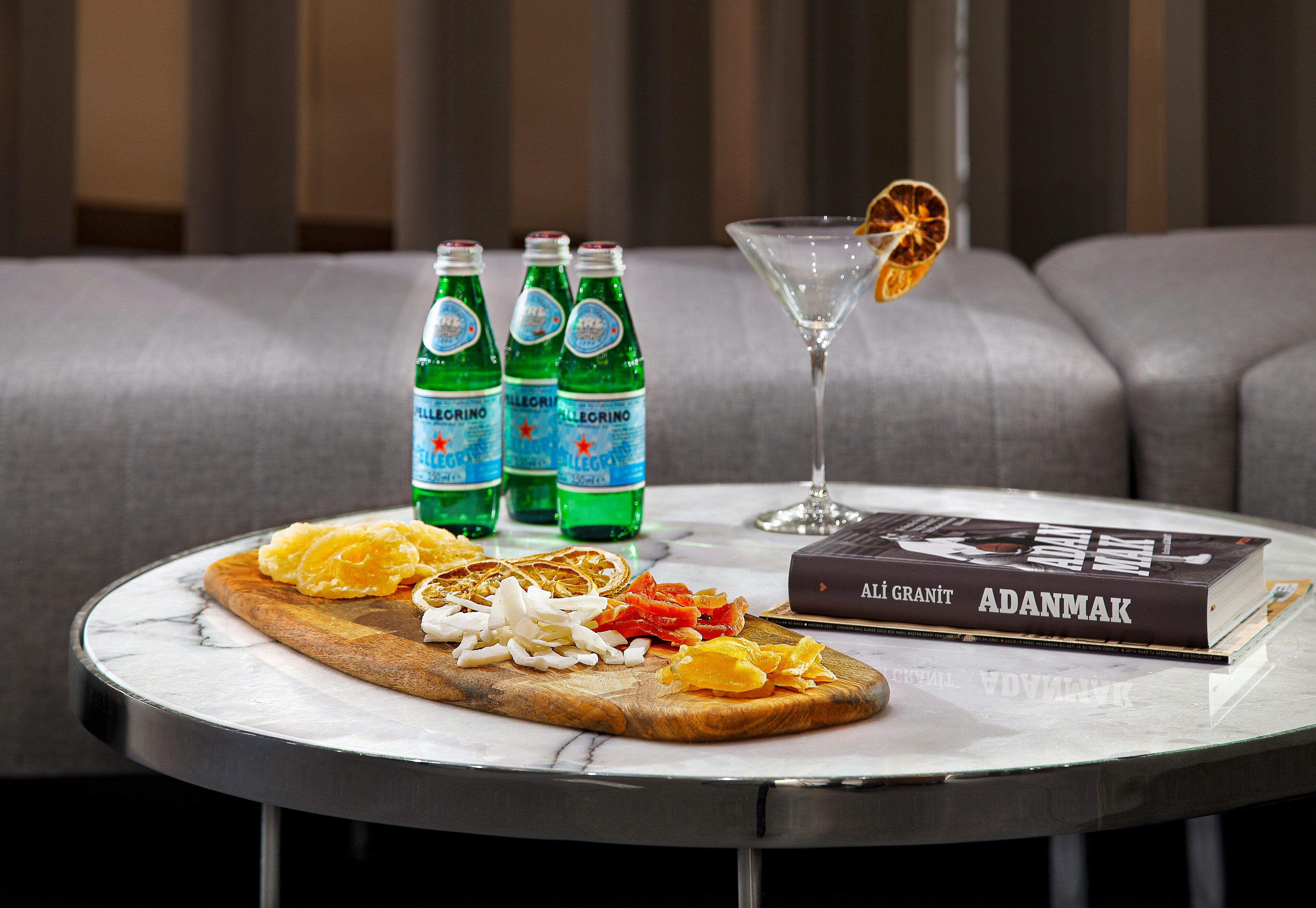 A round table with a marble top, a book titled 'ADANMAK', three bottles of 'Pellegrino' water, a martini glass with an orange slice, and a wooden board with sliced oranges and onions.