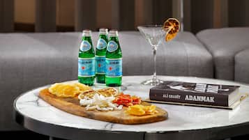 Una mesa redonda con tapa de mármol, un libro titulado 'ADANMAK', tres botellas de agua 'Pellegrino', una copa de martini con una rodaja de naranja, y una tabla de madera con naranjas y cebollas en rodajas.