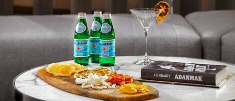 Una mesa redonda con tapa de mármol, un libro titulado 'ADANMAK', tres botellas de agua 'Pellegrino', una copa de martini con una rodaja de naranja, y una tabla de madera con naranjas y cebollas en rodajas.